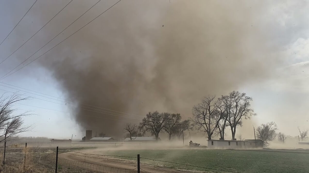 Large dust storm causing severe soil erosion across farmland with trees and buildings in background